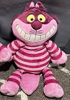 Photo of the Scentsy Disney Cheshire Cat plush showing the full body with pink and magenta stripes, smiling face, and detailed facial features including wide eyes and nose.