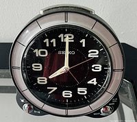 Front-facing image of a Seiko table clock showing black dial with light-up Arabic numerals, silver hands, and a silver-tone bezel. The clock face includes a second hand and control knobs are visible along the base for setting functions.