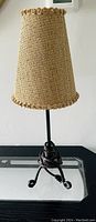 Full view of the table lamp showing woven beige shade and black rod iron legs on a clear glass surface.