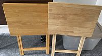 Photo showing two wooden TV trays standing upright with the tops facing the camera, revealing wood grain and folding leg structure.