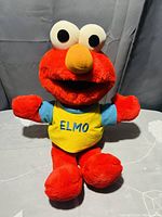 Front view of red Elmo plush toy with yellow shirt labeled 'ELMO'