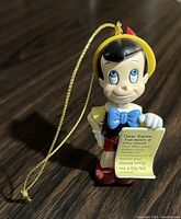 Single photo showing vintage Pinocchio figure Christmas tree ornament with a gold hanging string, standing on a wooden surface.