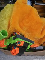 Orange and yellow large plush toy partially visible with green, black, and orange Nerf blaster in box.