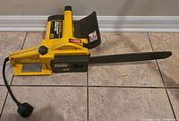 Side view of yellow and black McCulloch Electramac EM250 electric chainsaw lying on tiled floor, showing entire body and saw bar with electric cord attached.