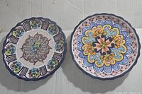 Pair of signed Talavera Mexican pottery plates with colorful floral and geometric patterns and scalloped edges, placed side by side.