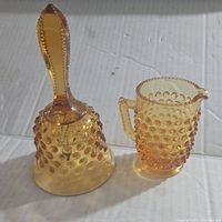 Amber hobnail glass bell and mini creamer pitcher displayed side by side on white surface, showing texture and shape of both items