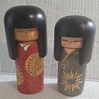Front view of two vintage Kokeshi dolls, showing red and green kimonos with floral motifs and classic black hair bob style.