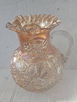 Full view of vintage carnival glass pitcher with ruffled top and handle, showing iridescent orange color and grape leaf pattern.
