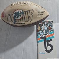 Overall view of the Miami Dolphins football with vintage logo and Dolphins 1975 Facts Guide booklet beside it.
