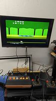 Atari Flashback 2 console powered on and connected to a TV displaying a classic Atari game screen with score and timer visible. Console with two joysticks and wired connections seen on a table.