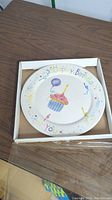 Top-down view of white ceramic birthday plate in box, showing cupcake with candle and balloon design and 'Happy Birthday To You' text around rim.