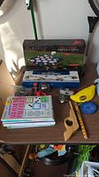 Photo of lot showing boxed Black Edition Rack 'Em Up Portable Pong game, Wooden Tabletop Cornhole box, puzzle books, and various toys on table.