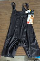 Top view of black Miracle Suit shapewear bodysuit with original tags attached, showing front body and straps.