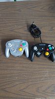 Two Nintendo GameCube controllers on a wooden surface. Silver WaveBird wireless controller on left and black wired controller on right.