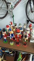 Photo showing 14 assorted wooden nutcracker figures arranged on a table beneath a bicycle wheel, featuring various colorful designs and styles.