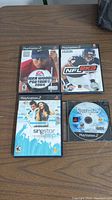 Four PlayStation 2 games on wooden surface: Tiger Woods PGA Tour 2004, NFL 2K3, SingStar Pop, and loose disc labeled Rom 2 Spy.