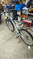 Full side and rear angle view of white and turquoise Rallye 18 speed bicycle showing frame, tires, gears, pedals, and handlebars in a garage setting.