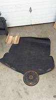 Black rubber trunk mat laid out on concrete floor with round rusty metal weight placed on top to hold it down, showing the mat's overall shape and condition.