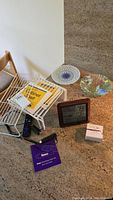 Overview of lot items including two fused art glass plates, digital weather station, white cabinet racks, Roku remote and streaming stick, and quick start guide booklet on carpeted floor.