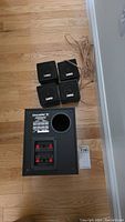 Five piece sound system components including four satellite speakers and one subwoofer on wood floor with visible speaker wire.