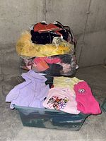 Stacked clear bin containing pastel colored children's clothing and a large bag with more clothing and dress-up items on top in a basement setting.