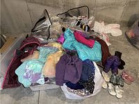 Wide view of children's clothing pile on concrete floor including coats, dresses, shirts, and costumes packed loosely in bags