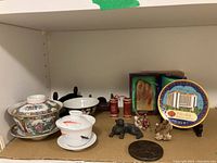 Shelf displaying assorted small figurines, decorative plates, carved jade collectible, and ceramic rice bowls with lids and stands.