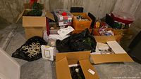 Wide shot showing multiple boxes and bags containing assorted Christmas decorations including tree ornaments, wreaths, and boxes stored in a basement space.