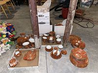 Overview of the orange cloisonné dishware including bowls, teacups, plates and serving dishes arranged on floor near wooden posts in basement.