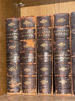 Books arranged on bookshelf showing worn black leather spines with gold embossed titles for 'Barnaby Rudge, Edwin Drood,' 'Bleak House,' 'Child's England, Esther,' and 'Christmas Books, Reprinted Pieces.' Significant wear and peeling on spines.