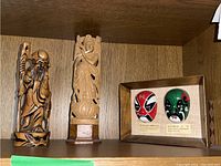 Two Asian styled wood figurines and one framed wall art with masks displayed on a wood shelf.