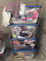 Four clear plastic tubs stacked, filled with assorted infant and toddler clothing items such as sleepers, jackets, dresses, indicating volume of lot.