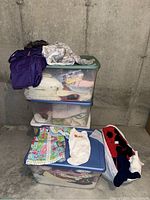Photo showing stacked plastic bins filled with infant and toddler clothing including winter coats and blankets. Some items like jackets and sweaters are folded on bin tops.