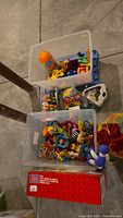 Photo showing two transparent plastic bins filled with assorted infant toys including rattles, teethers, and small toys, taken on concrete floor.