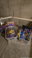 Three plastic storage bins containing multiple LEGO boxes and loose parts in a basement corner
