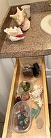 Overhead view of open drawer showing plastic container with polished stones, carved stones, raw rock, and chips of various sizes with ceramic items on countertop