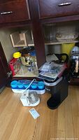 Photo of cabinet with single cup Keurig coffee maker, Black & Decker chopper, plastic containers and other small kitchen items.