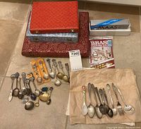 Overall assortment of silverplated spoons, souvenir spoons, forks, serving utensils and cleaning aids laid out on floor next to three decorative storage boxes and various packaging