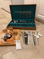 Wooden cutting board with wooden sushi accessories and artificial sushi. Assorted metal, wood, ceramic chopsticks displayed in front. Wooden case with stainless steel flatware and long skewers at back.