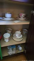 Wide view of wooden cabinet interior showing multiple teacup sets and saucers on three shelves with varied patterns and sizes