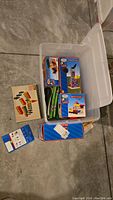 Plastic container holding Thomas The Tank Engine boxed track parts and accessories along with a vintage Brio wooden building set box, placed on cement floor.