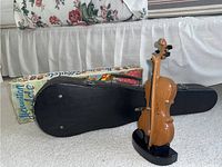 Wooden violin standing upright beside black hard case and vintage ukulele box on floor.