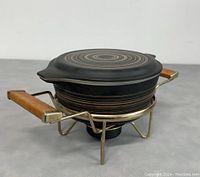 View of covered casserole dish atop candle warmer stand, showing circular pattern and wooden side handles