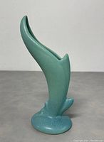 Front angled view of the turquoise-blue-green matte glazed pottery vase shaped like a Bird of Paradise flower, showing its curved top opening and rounded base on a neutral grey surface.