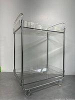 Front angled view showing clear lucite top and bottom shelves, chrome frame, and caster wheels.
