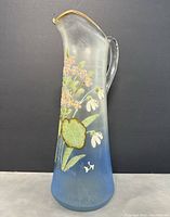 Front view of the tall slender glass jug, showing blue frosted base and hand painted pink and white flowers with green leaves, designed in a naturalistic style.