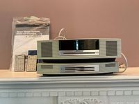 Front view of stacked Bose Wave Music System main unit atop multi-CD changer with two remotes and sealed manuals behind
