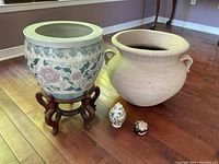 Two large planters side by side on floor, one ceramic on wooden stand and one large clay pot. Two small decorative containers in front