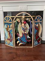 Full front view of the three-panel metal folding fireplace screen with painted angels and hanging bulbs in front of a fireplace.