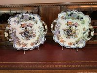 Two ornamental square-shaped decorative plates with handles, featuring delicate floral designs with gold accents, set on clear display stands.
